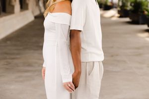 Two people in all-white summer outfits hold hands while standing close together in an outdoor covered walkway.