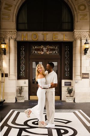 A couple embraces outside an elegant hotel entrance with ornate architectural details and black and white geometric flooring.