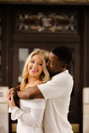 Romantic couple embraces in an elegant indoor setting wearing matching white outfits.