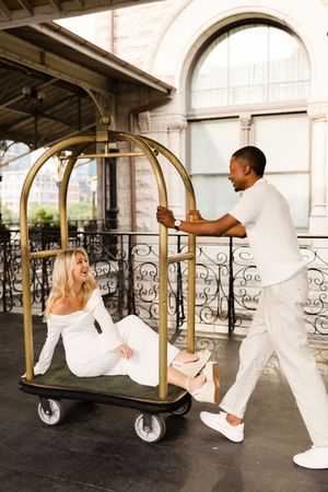 Stylish couple in all white outfits having fun with luxury hotel luggage cart on an elegant covered balcony.