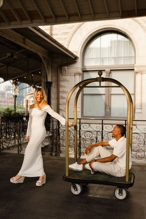 Playful couple in white outfits creates dynamic poses on a vintage luggage cart under arched architecture.
