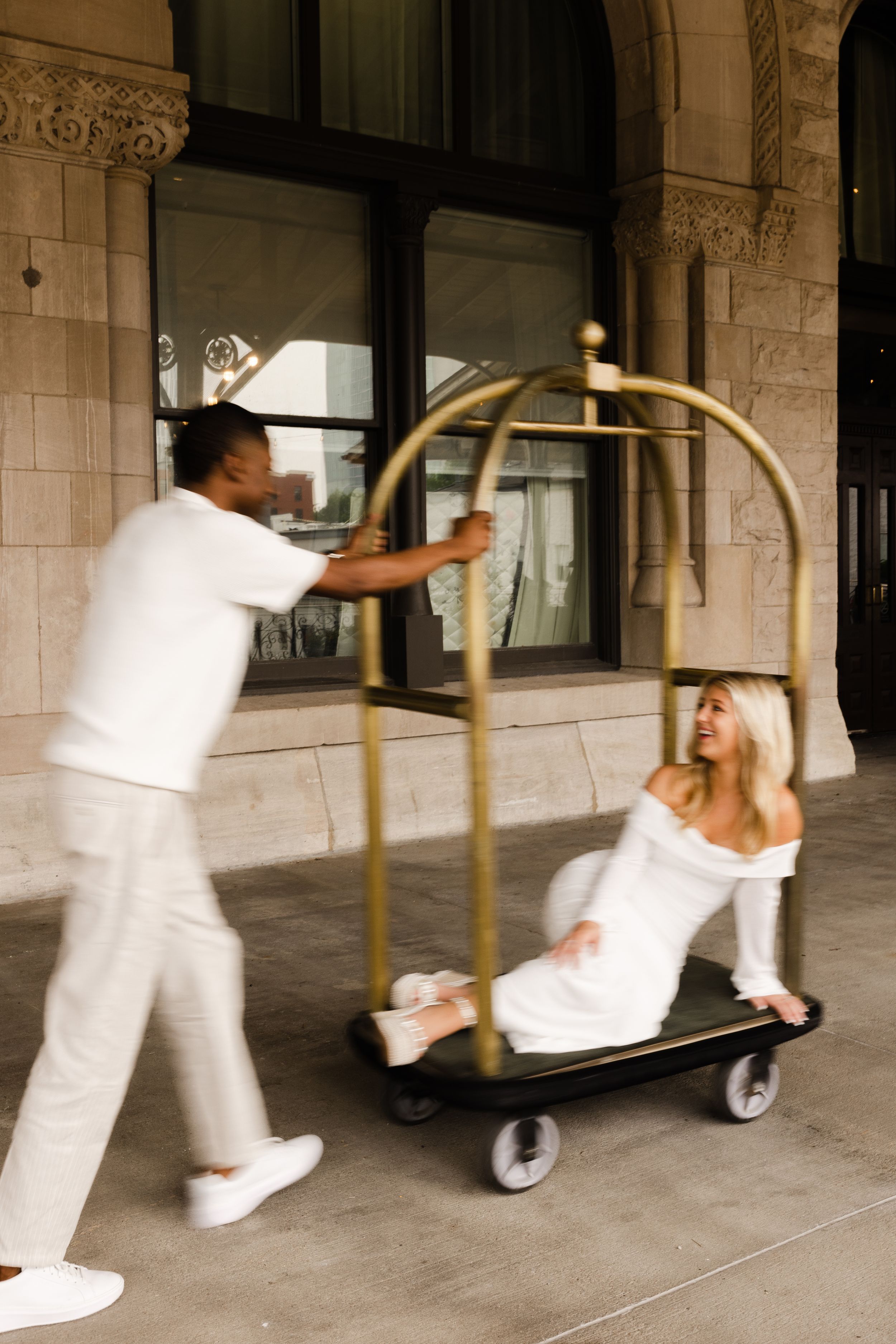 Person in white uniform pushing gold luggage cart with passenger in white dress at elegant hotel entrance.