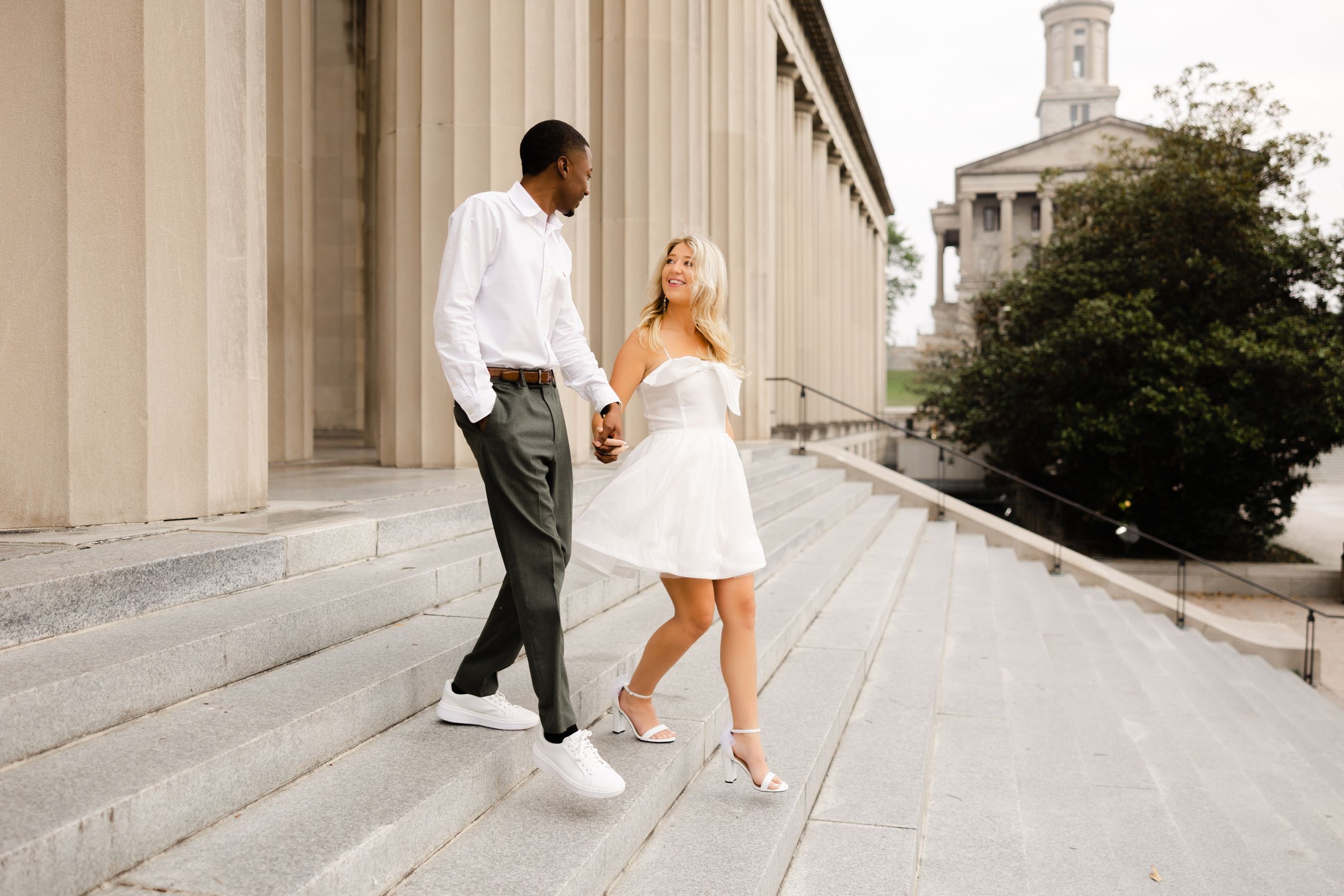 A couple walks together on grand stone steps outside a stately building with columns on a sunny day.