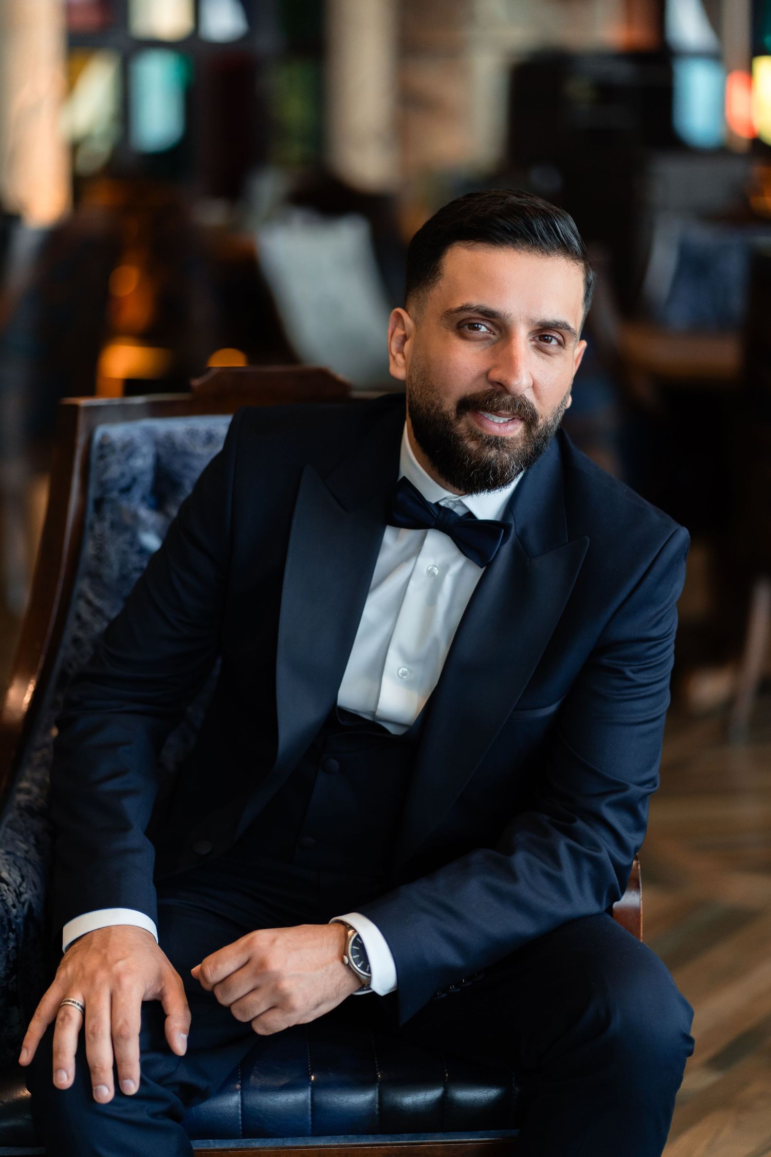 Man in formal black tuxedo and bowtie sits elegantly on leather chair in dimly lit setting.