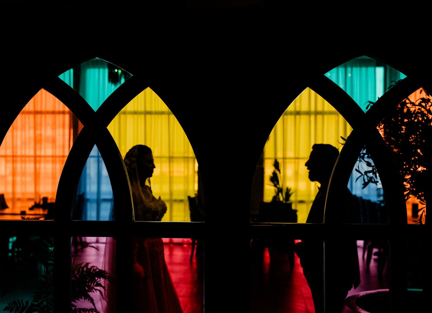 Silhouetted figures stand against colorful stained glass church windows glowing with red, yellow, and turquoise light.