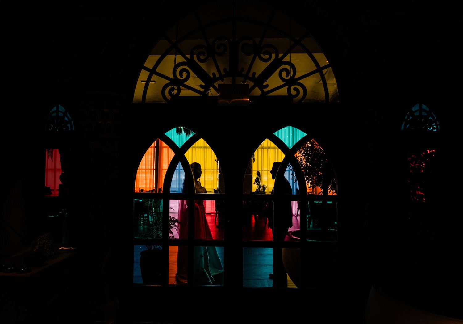 Silhouettes stand behind colorful stained glass church windows glowing with yellow and orange light in darkness.