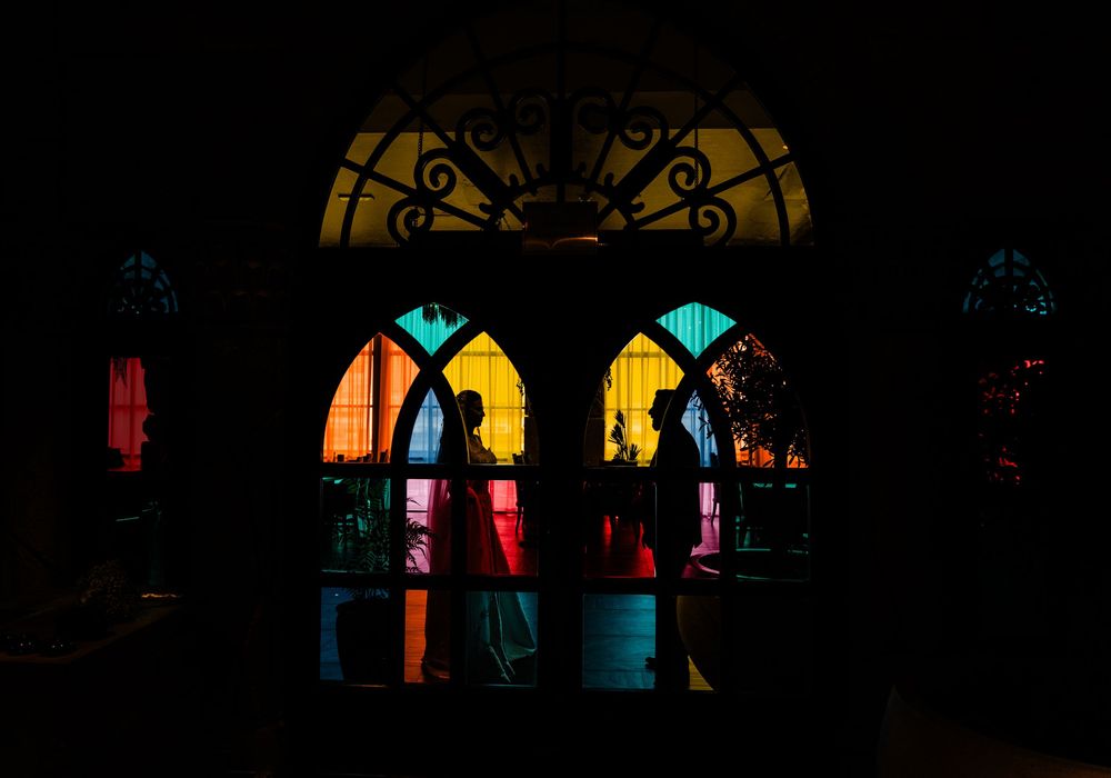Silhouettes stand behind colorful stained glass church windows glowing with yellow and orange light in darkness.
