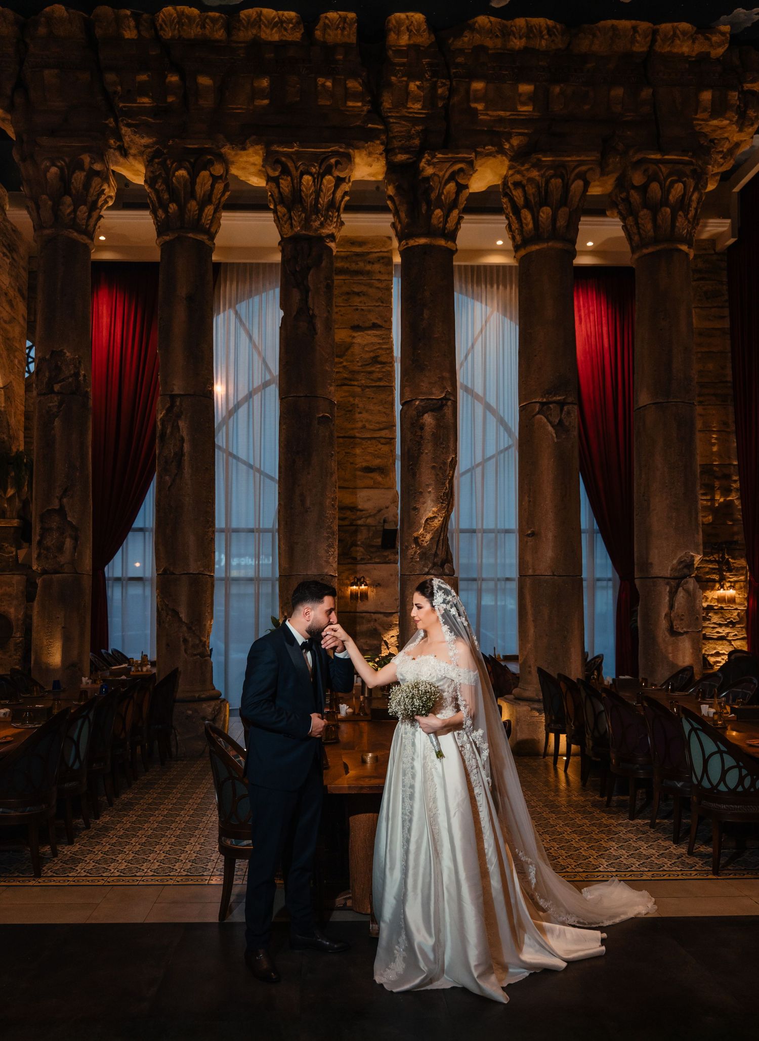 Elegant wedding ceremony in a historic columned venue with dramatic lighting and red velvet curtains.