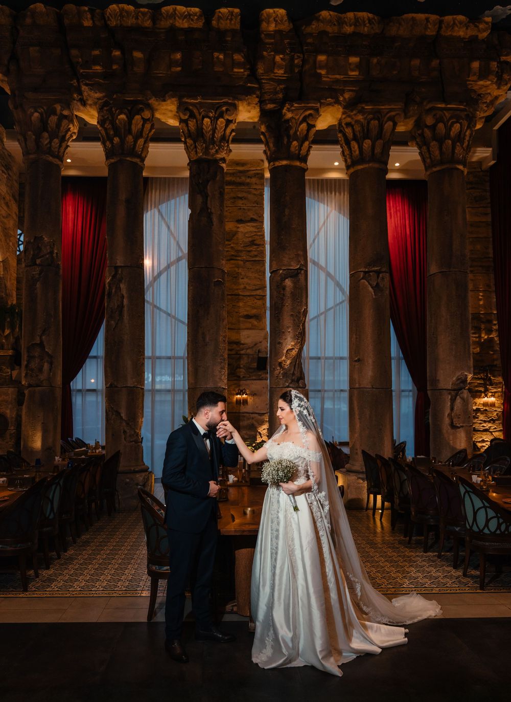 Elegant wedding ceremony in a historic columned venue with dramatic lighting and red velvet curtains.