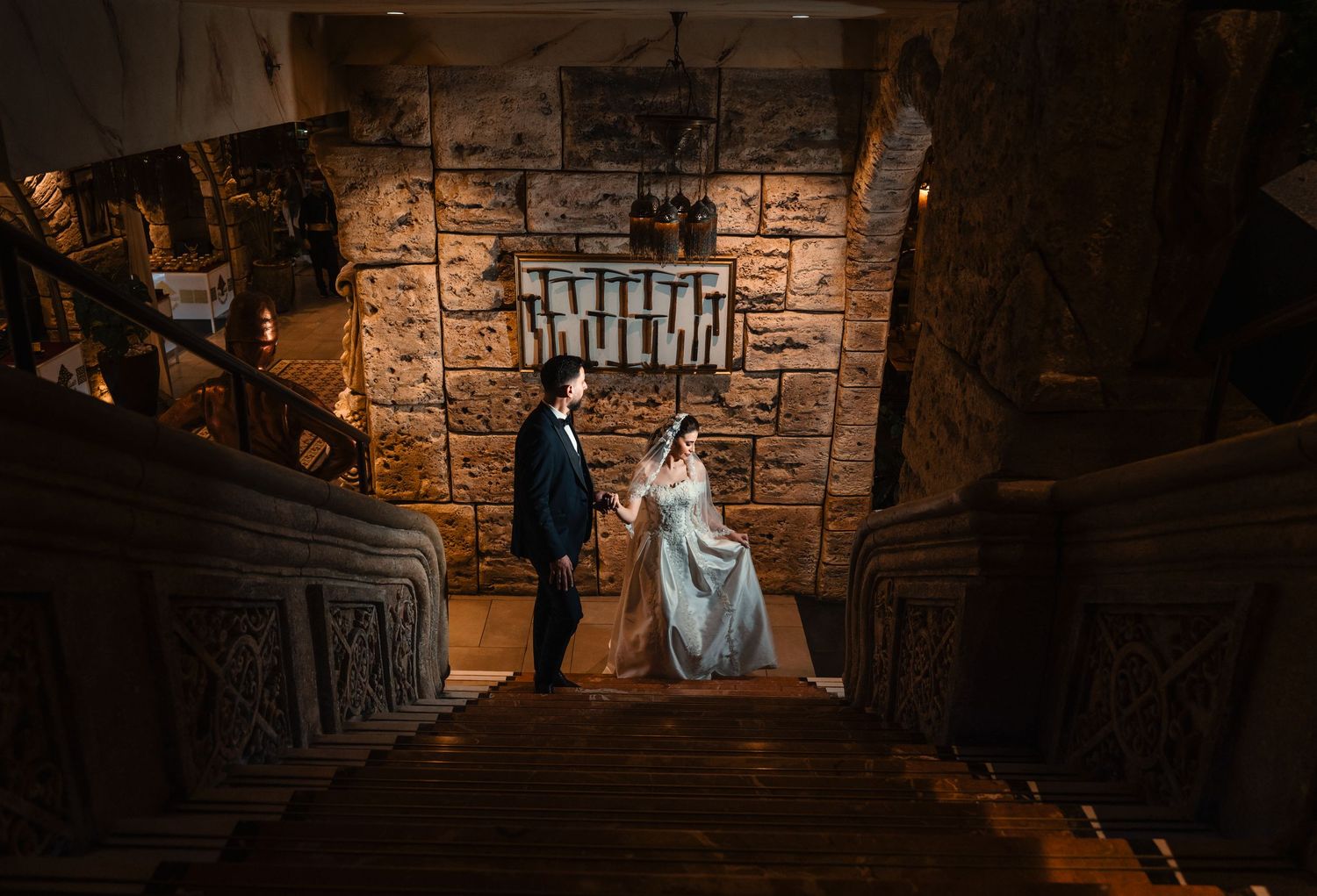 A couple in formalwear stands in a dramatic stone stairwell illuminated by warm lighting in a vintage-style setting.