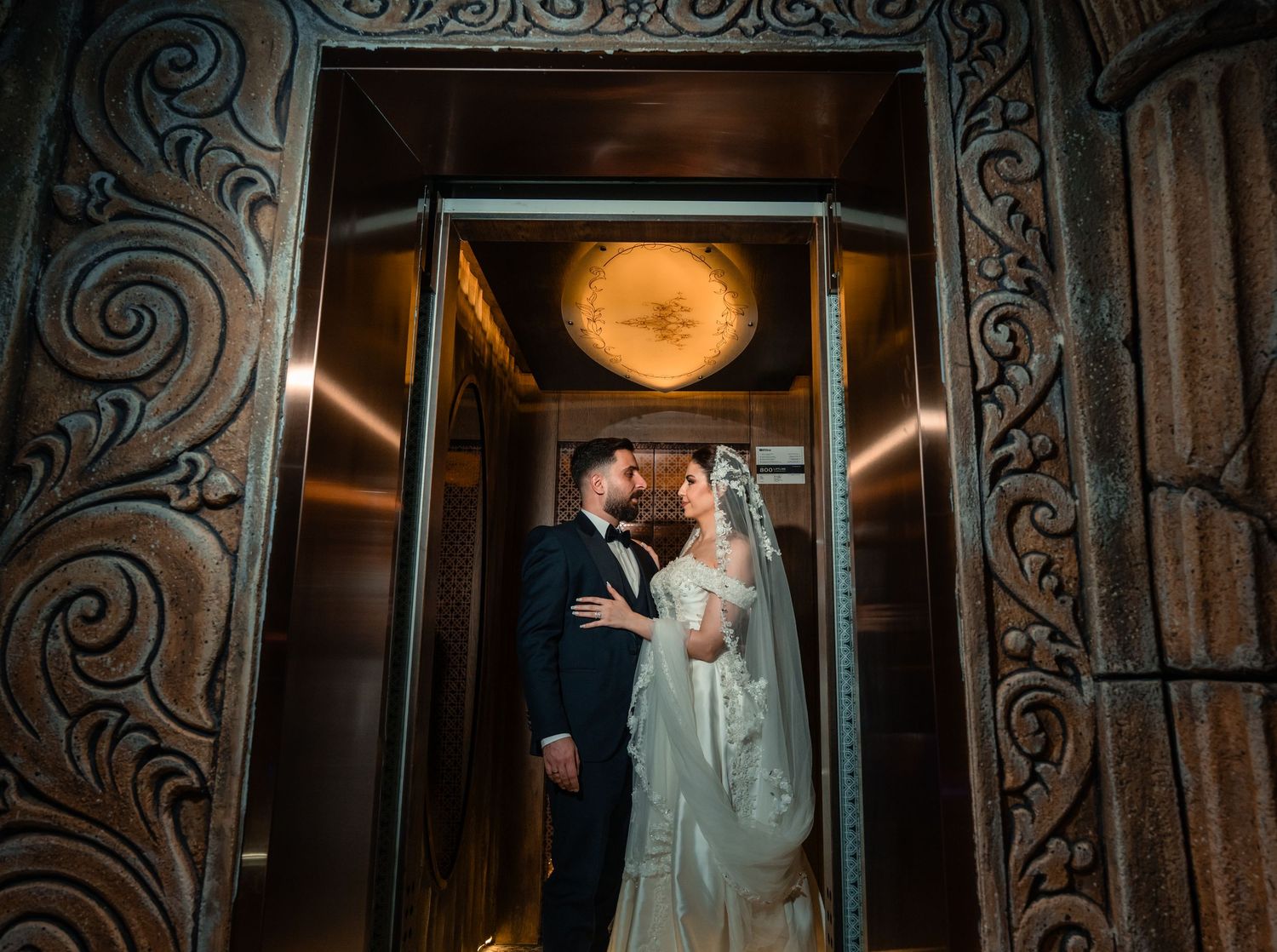 Couple embraces in ornately decorated vintage elevator with warm glowing light and art nouveau swirl details.