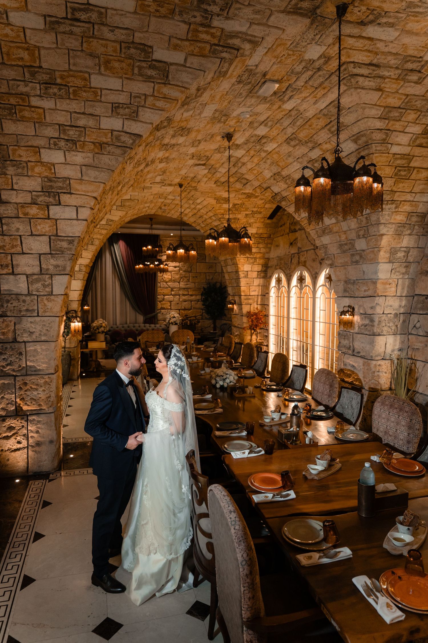 Castle dining room with stone arched ceiling, ornate chandeliers and long wooden table set for an elegant wedding reception.
