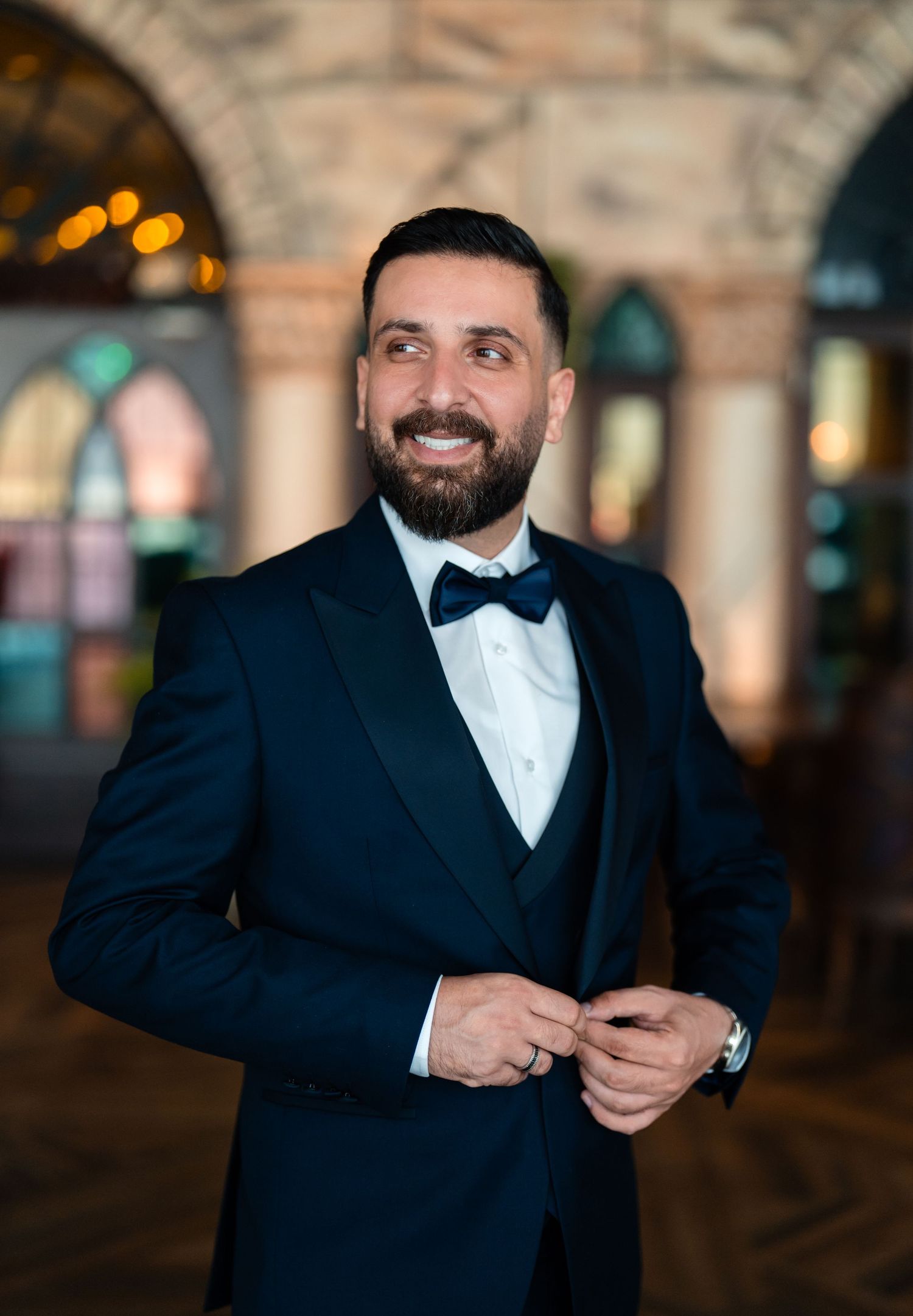 Elegant groom in black tuxedo and bowtie smiling while standing in venue with arched architecture and glowing lights.