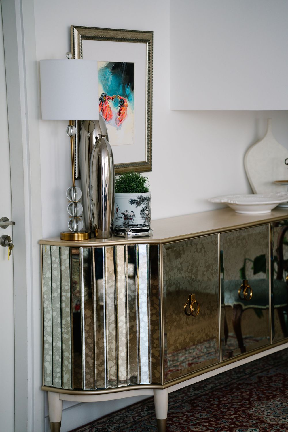Mirrored sideboard cabinet with chrome lamp and framed artwork in a modern interior setting.