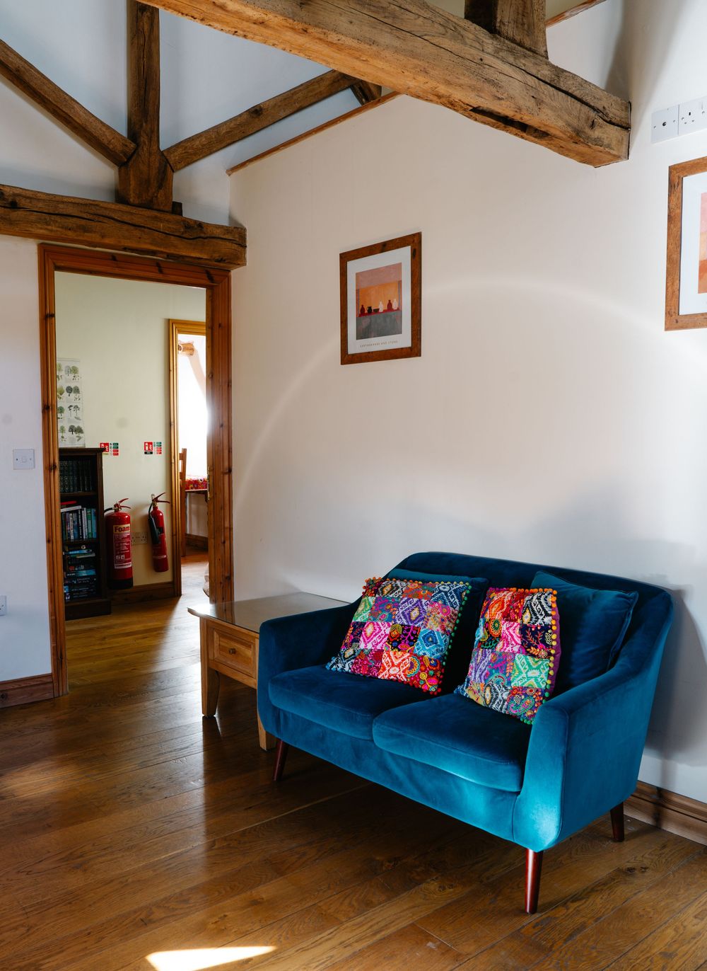 Blue velvet loveseat with floral cushions beneath exposed wooden beams in a bright interior space.