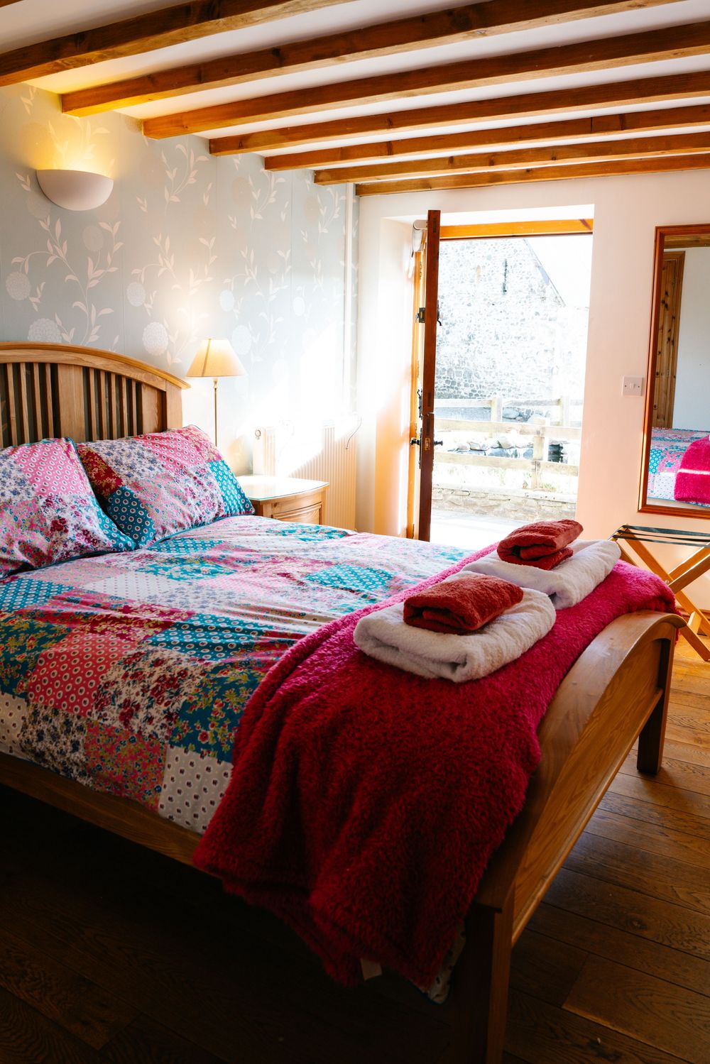 Cozy bedroom with colorful patchwork quilt, exposed ceiling beams, and bright natural lighting.