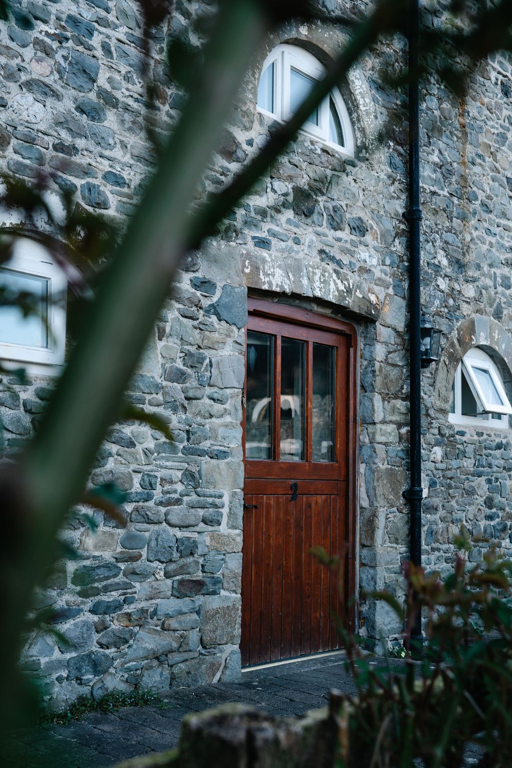 Weathered wooden door set in an old stone wall with small windows viewed through branches.