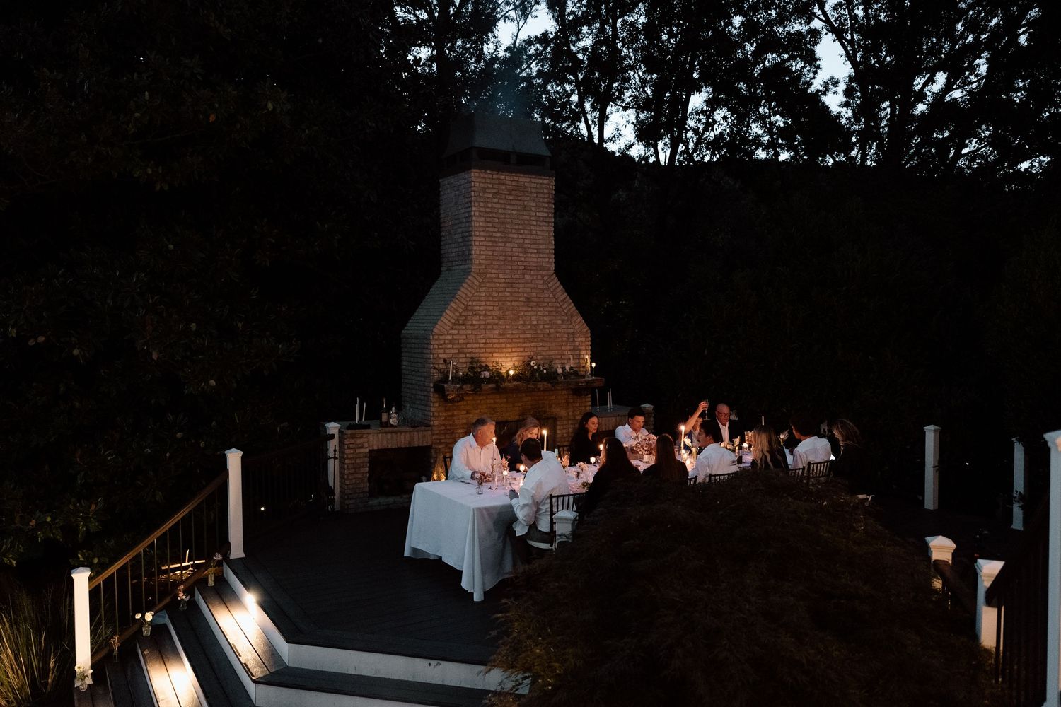 An intimate outdoor wedding ceremony takes place under trees on a grassy lawn with white chairs for guests.