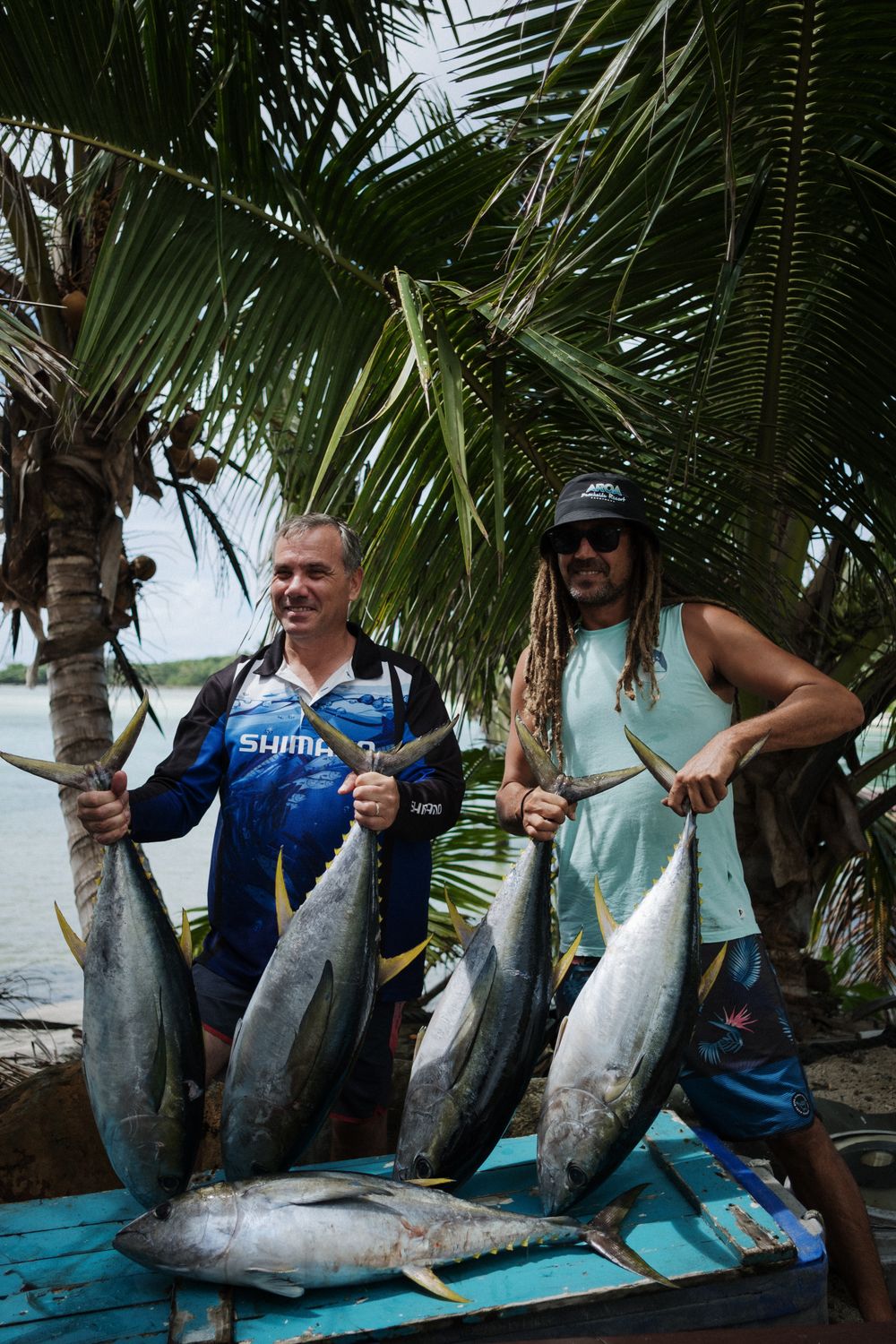 Two fishermen display their impressive catch of large tuna fish while standing near palm trees on a blue deck by the water.