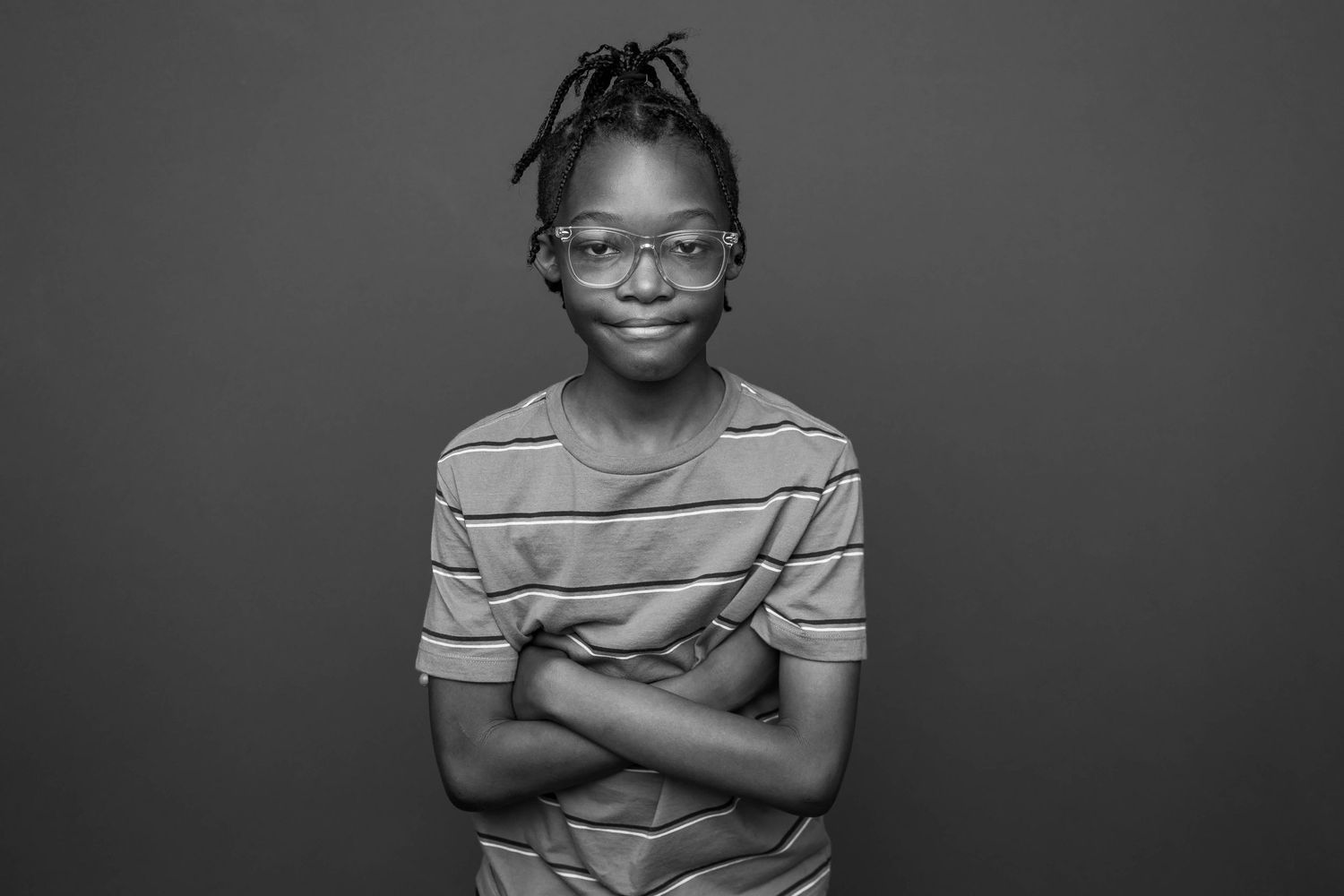 A young student wearing glasses and a striped shirt smiles while standing against a gray background in black and white.