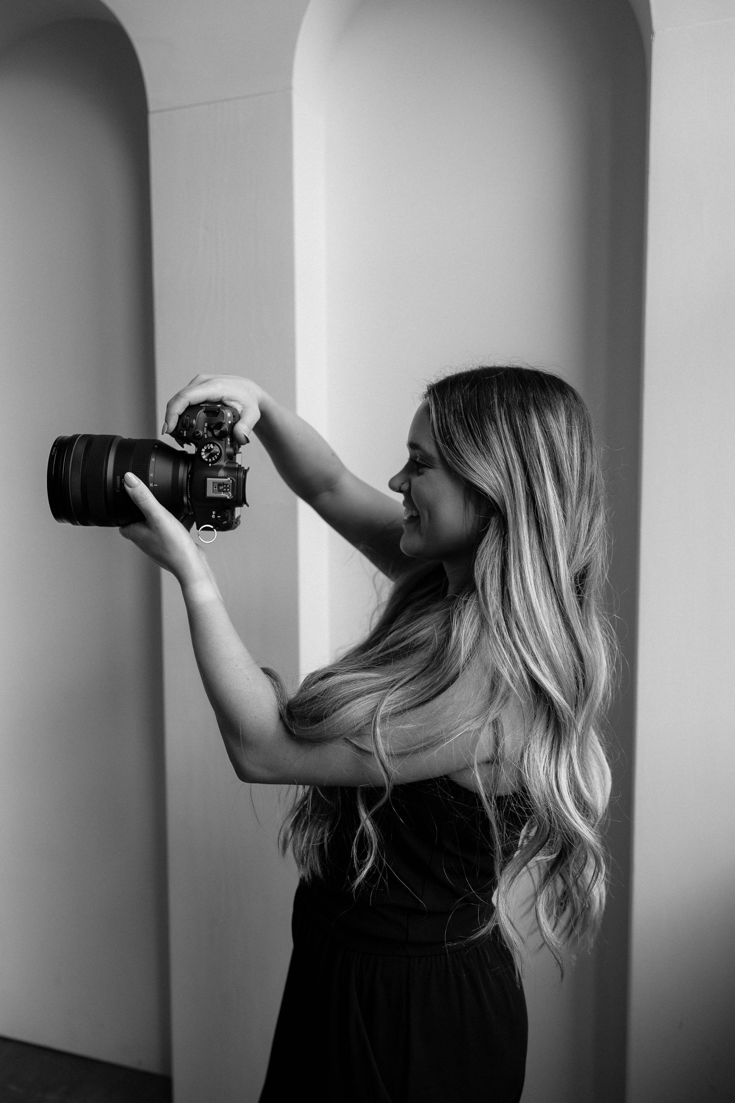 Black and white portrait of photographer in elegant dress holding professional camera up to take photo in arched hallway.