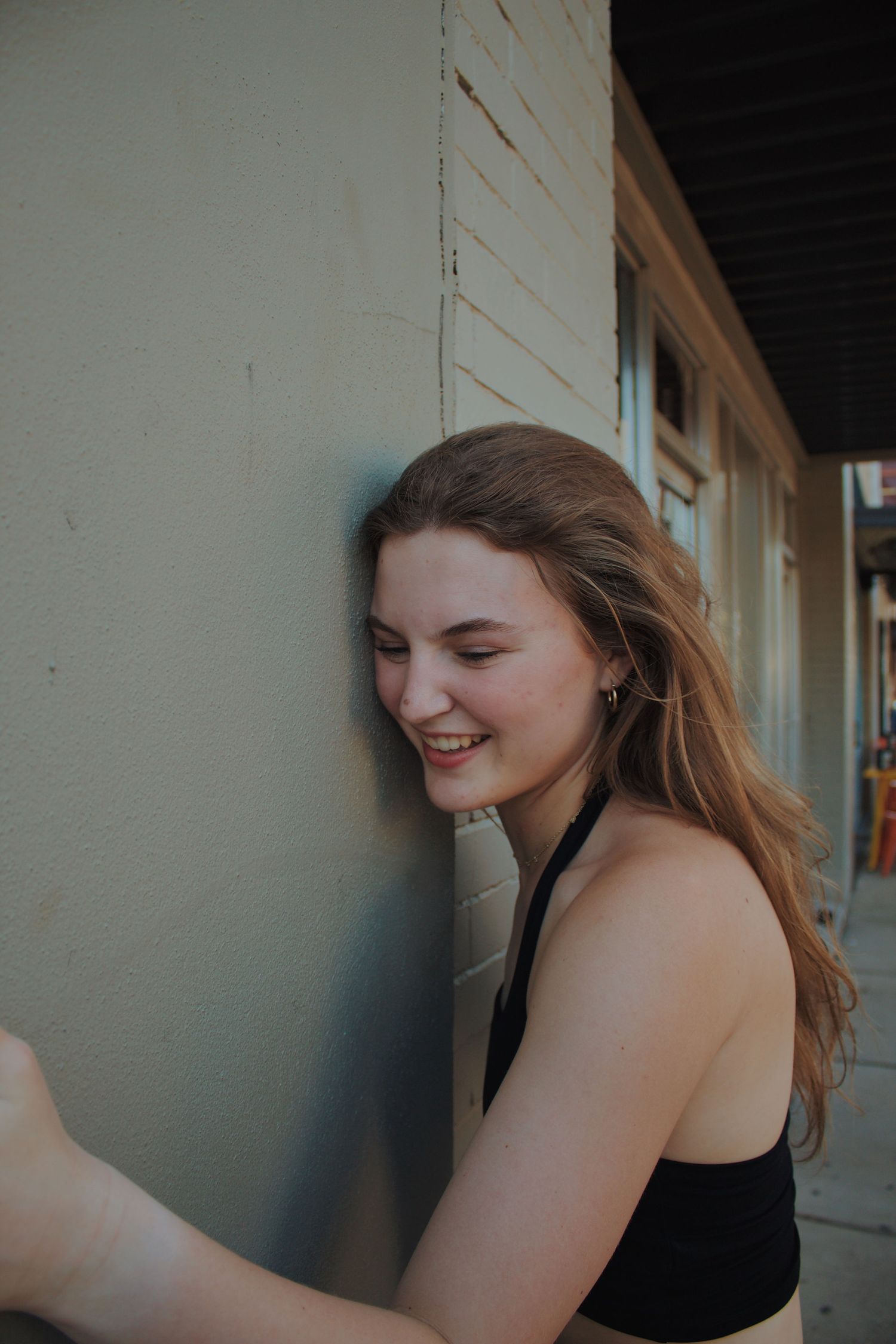 A profile portrait of a joyful person with flowing brown hair laughing while leaning against a wall outdoors.