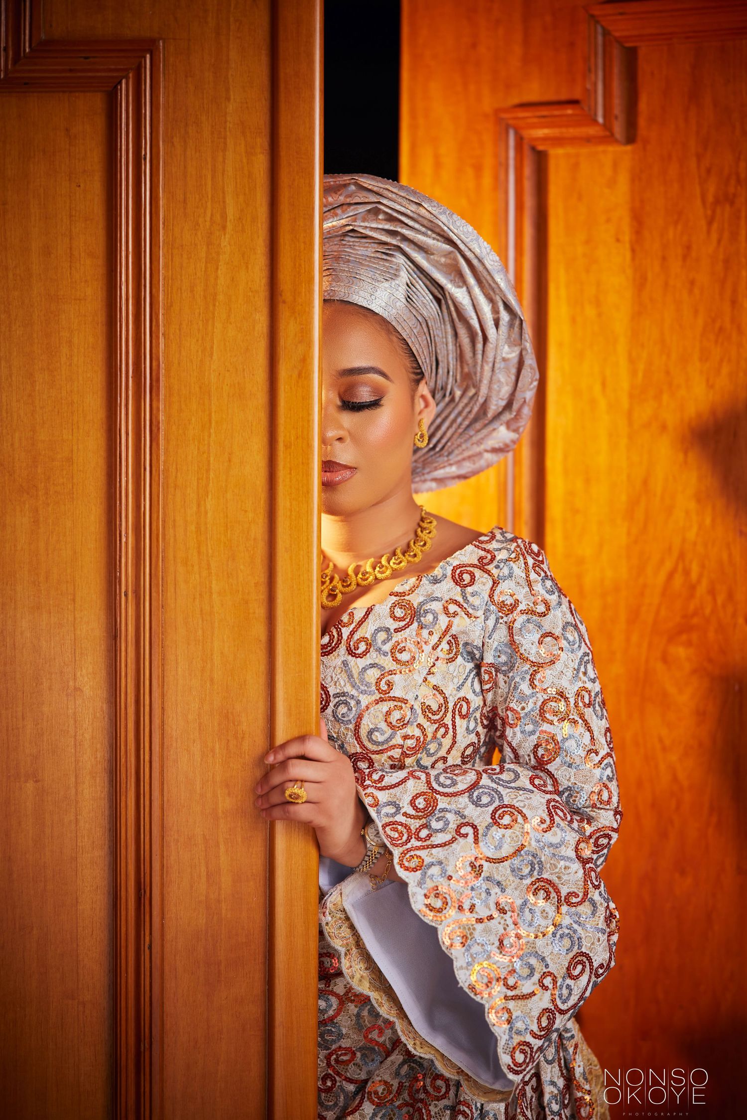 Portrait of person in traditional gele headwrap and patterned dress standing by wooden doors.