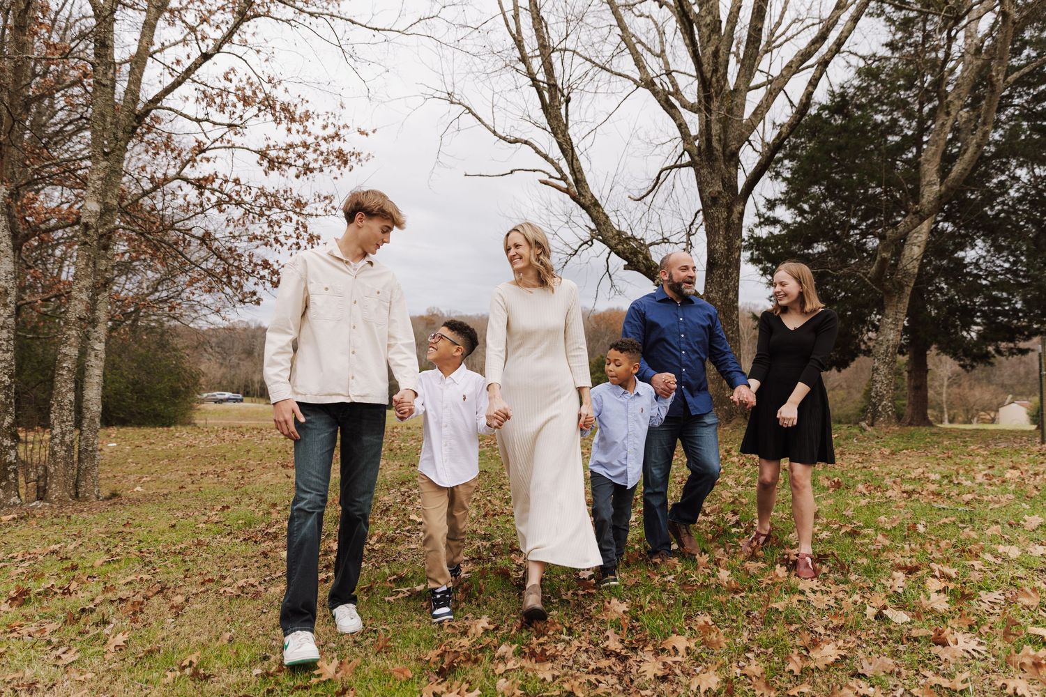 A family group walks together on a grassy hillside holding hands during autumn with bare trees in background.