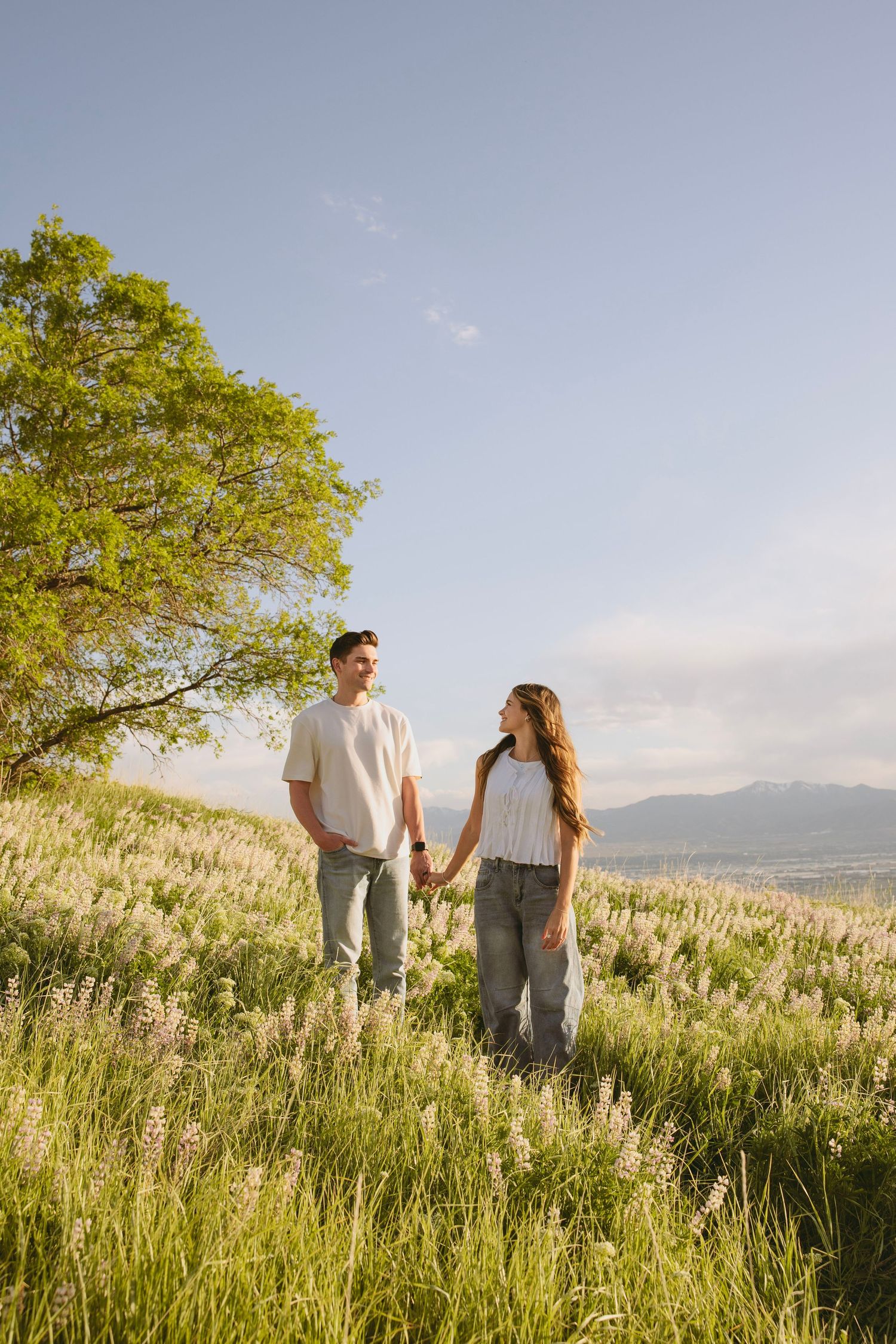 A couple walks hand in hand through a sunlit meadow with mountains in the background and a tree overhead.