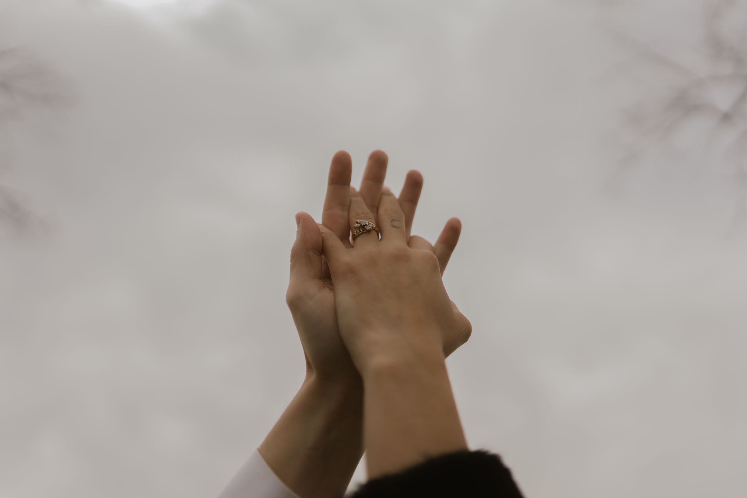 Close-up of hands raised against cloudy sky showing engagement ring.