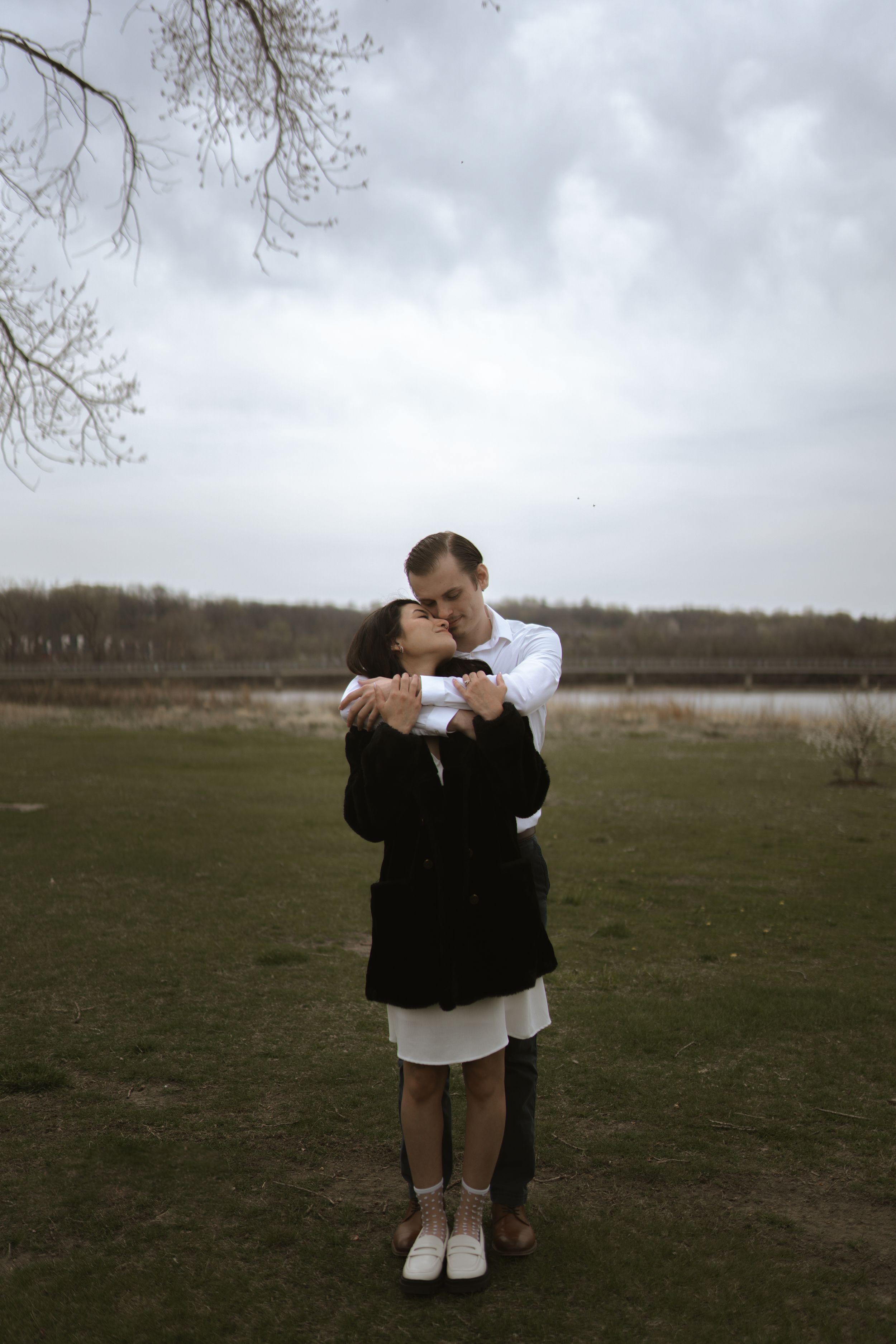 A couple shares an intimate moment standing in an open field near a lake.