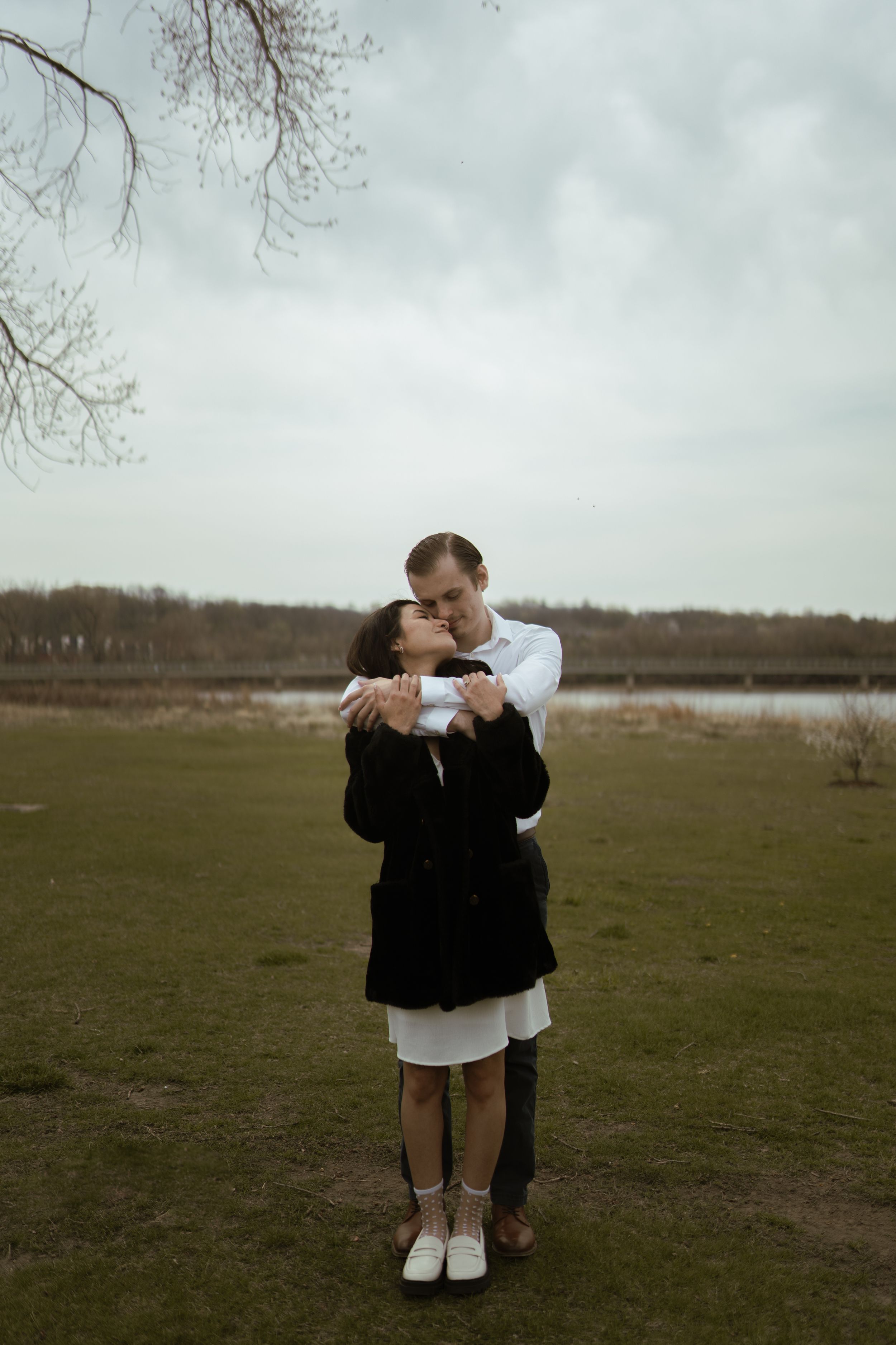 Two people embrace while standing in a misty field with bare trees in the background.