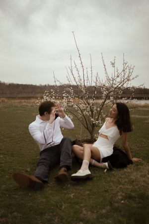 Couple lounges on grass sharing drinks near blooming tree.
