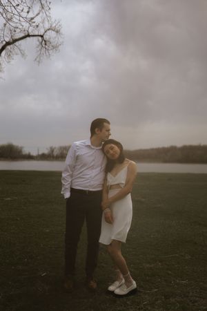 A romantic couple embraces on a cloudy day in an open field with bare trees in the background.