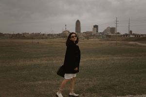 A person in a black jacket poses in front of an urban skyline on an overcast day.