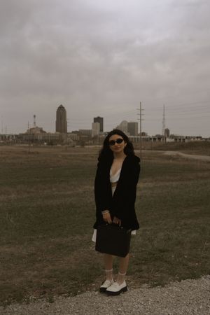 A fashionable person in black poses with a handbag against an industrial cityscape.