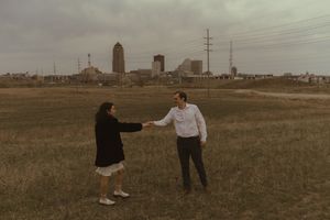 A couple dances playfully in a field with urban buildings in the distance.
