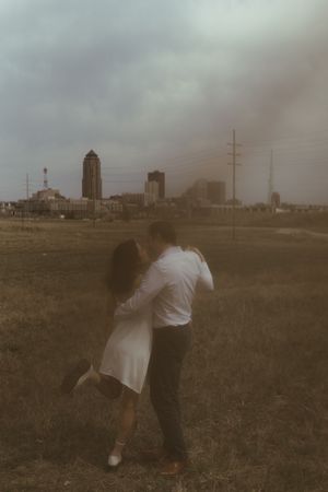 A couple in white and black attire embrace in front of a cityscape on an overcast day.