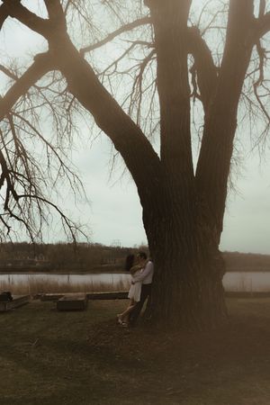 A moody sepia-toned photo of a couple embracing beside a large tree trunk near a lake at dusk.