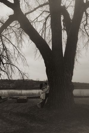 Black and white photo of two people standing against a massive tree trunk with a lake in the background.