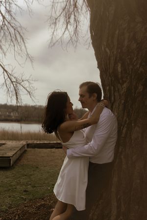 A romantic moment captured between a couple in white clothing embracing against a large tree trunk.