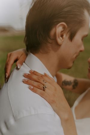 Close-up shot showing hands with an engagement ring resting on a white-clothed shoulder.