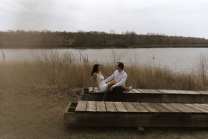 A couple sits together on a wooden dock by a serene lake surrounded by bare trees in winter.
