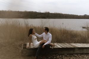 Couple in white clothing sitting together on a wooden dock by a serene lake.