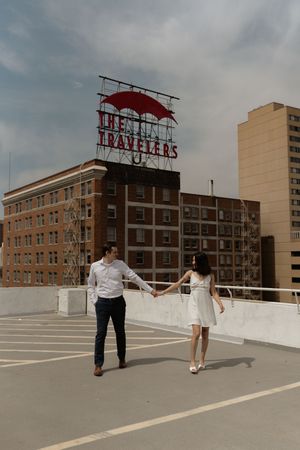 A couple holds hands while walking on a rooftop with the Travelers Insurance building behind them.
