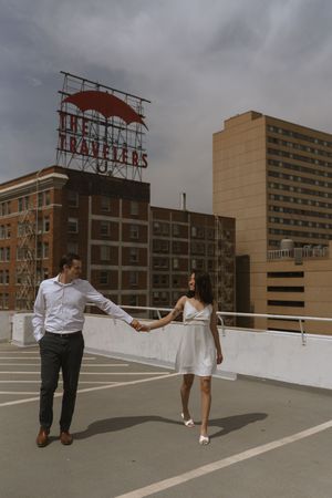 A couple in coordinated outfits holds hands on a downtown rooftop with dramatic sky behind them.