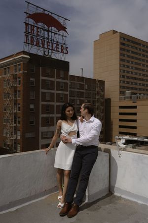 A couple sits together on a concrete ledge of a rooftop with urban buildings in the background.