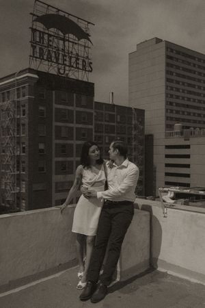 Couple dressed in contrasting white and dark outfits pose casually on a rooftop wall.