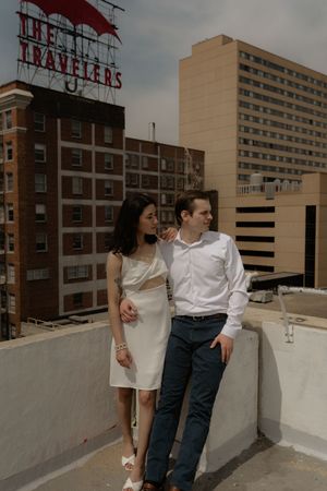 Two people share an embrace while sitting on a rooftop wall with city buildings behind them.
