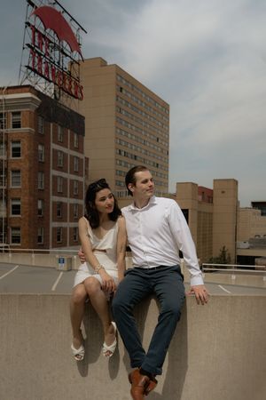 Two people sit casually on a concrete ledge overlooking city buildings on a cloudy day.